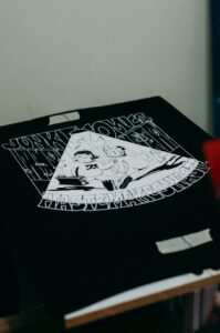 A person reviews digital artwork files on a laptop alongside printed T-shirt samples on a well-lit table.