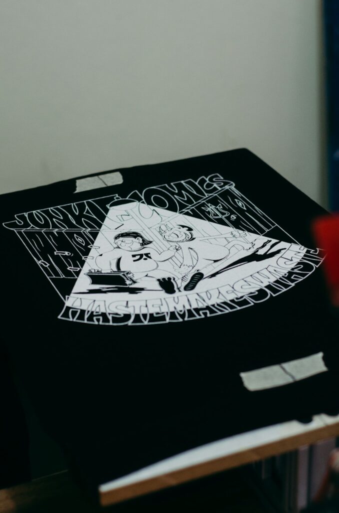 A person reviews digital artwork files on a laptop alongside printed T-shirt samples on a well-lit table.