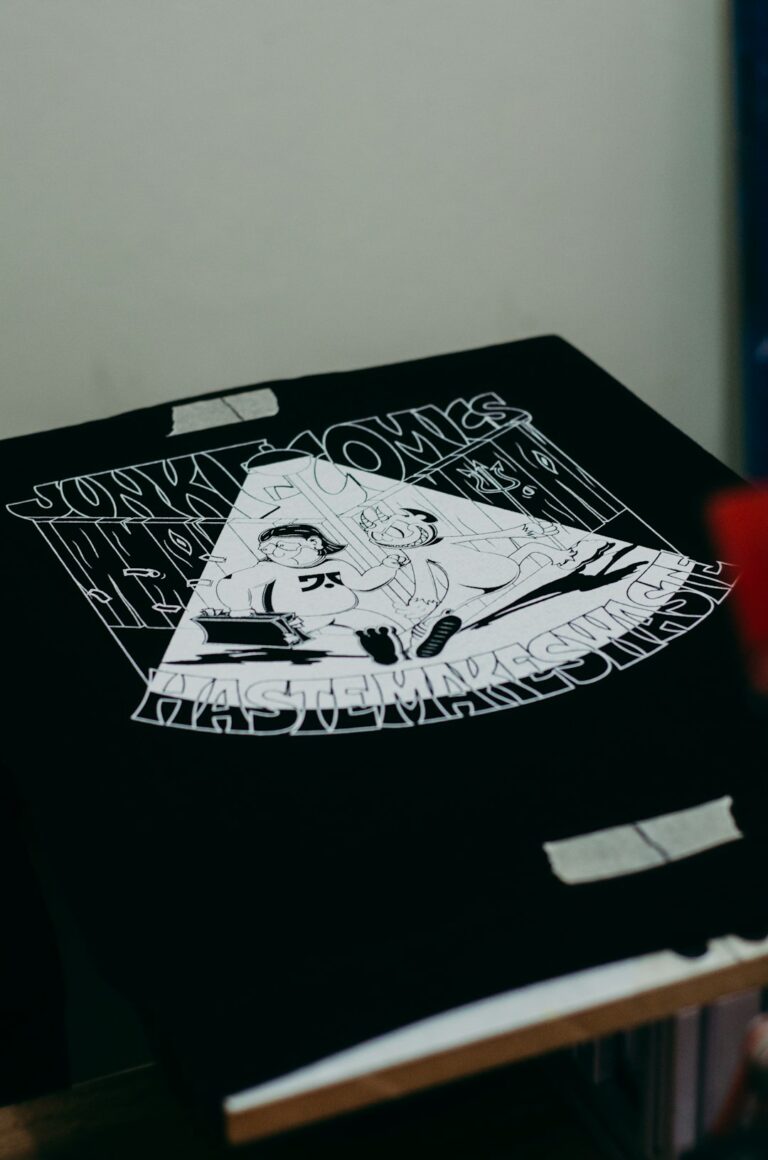 A person reviews digital artwork files on a laptop alongside printed T-shirt samples on a well-lit table.