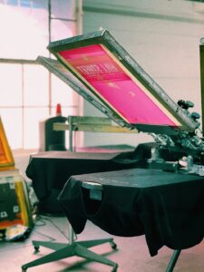 Close-up of hands inspecting a freshly printed t-shirt beside a printing press and digital printer.