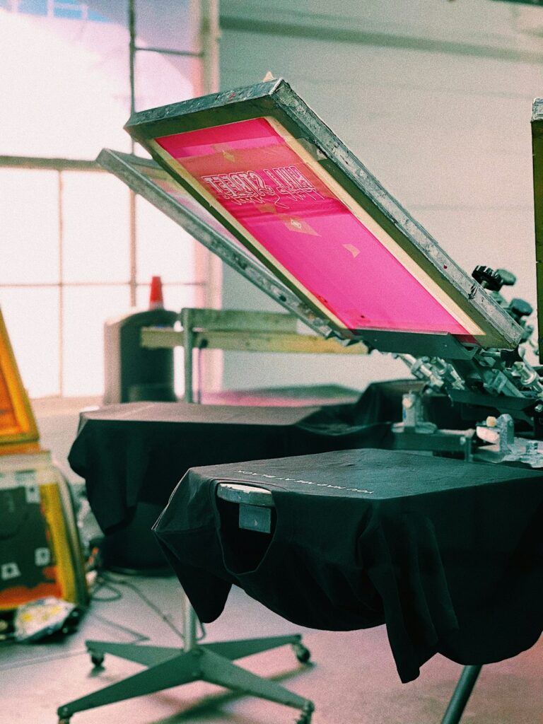 Close-up of hands inspecting a freshly printed t-shirt beside a printing press and digital printer.
